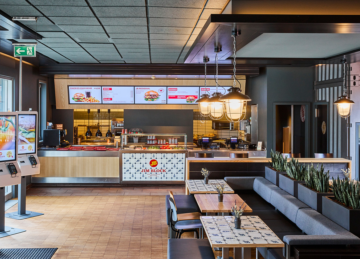 Jim Block Othmarschen Innenansicht Innenansicht des Jim Block Restaurants in Othmarschen mit modernen Sitzbereichen und digitaler Menütafel über dem Bestellcounter.